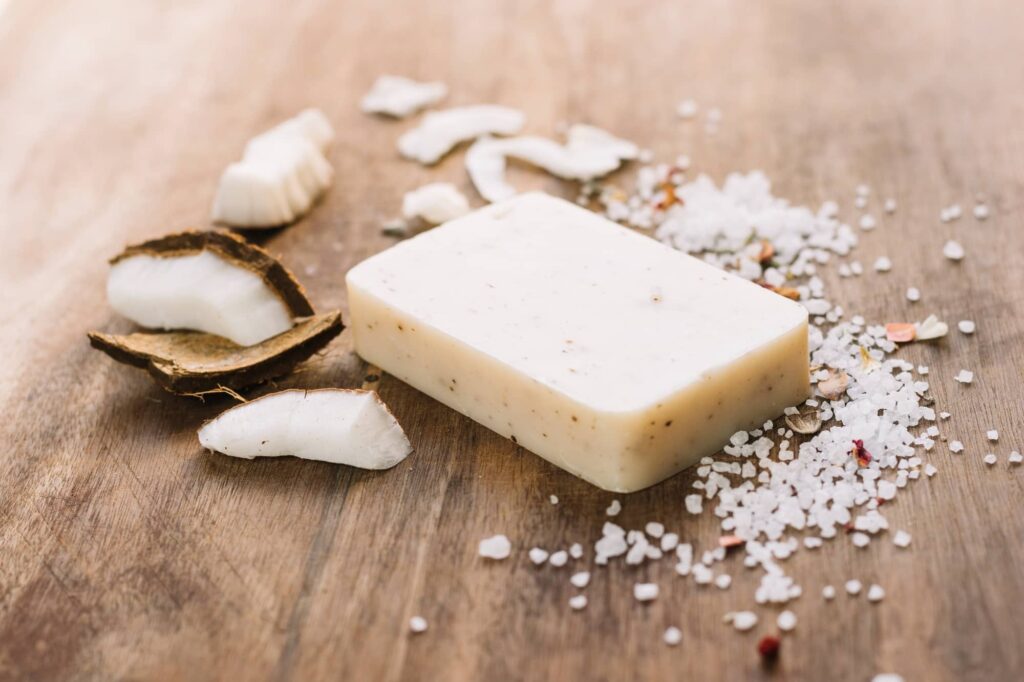 Immagine che mostra un panetto di balsamo solido per capelli di colore chiaro su un piano in legno. Il panetto sembra essere fatto con ingredienti naturali, forse all'olio di cocco, come suggeriscono i pezzi di cocco fresco sparsi intorno. C'è anche una sparsa di sale grosso e petali, che aggiungono un tocco di spa e benessere all'ambiente. La combinazione di questi elementi trasmette un senso di cura dei capelli naturale e organica. Il legno su cui poggiano il panetto e gli altri elementi aggiunge un senso di calore e semplicità all'immagine.