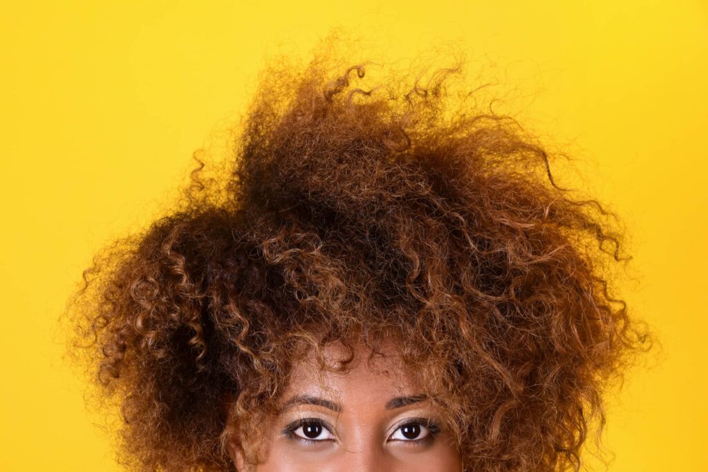 Inquadratura dal naso in su di una ragazza di colore con capelli ricci di media lunghezza molto crespi. Probabilmente si sta chiedendo quali siano i metodi per ridurre il crespo più adatti a lei.