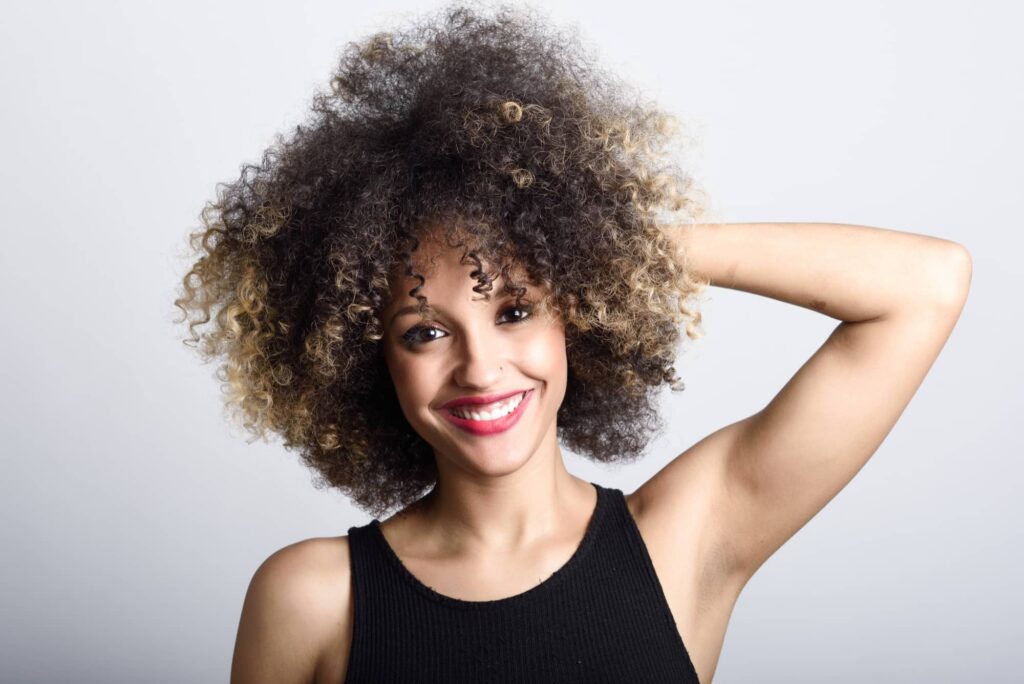 Ragazza sorridente con capelli ricci medio-lunghi molto crespi che si gratta dietro la testa in un gesto imbarazzato, probabilmente chiedendosi quali sono i migliori metodi per ridurre il crespo nei capelli ricci.
