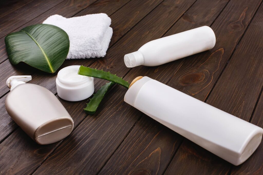 Immagine che mostra una serie di prodotti per la cura del corpo e dei capelli su un tavolo di legno scuro. C'è un asciugamano bianco piegato ordinatamente accanto a una foglia di pianta verde. Inoltre, sono presenti diverse bottiglie di plastica bianca che potrebbero contenere shampoo, balsamo o lozione. Una delle bottiglie ha un erogatore a pompa. Vicino a queste bottiglie, c'è un barattolo di crema con il coperchio aperto e un pezzo di aloe vera, noto per le sue proprietà idratanti e curative. Si tratta certamente dei migliori prodotti per capelli secchi.