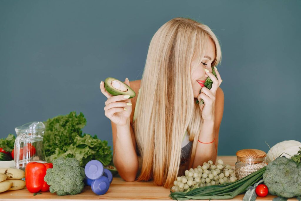 Una ragazza con lunghi capelli biondi che sorride mentre annusa delle verdure. Appoggia i gomiti su un tavolo di legno pieno di cibi salutari particolarmente indicati in un'alimentazione che mira a migliorare la salute dei capelli.