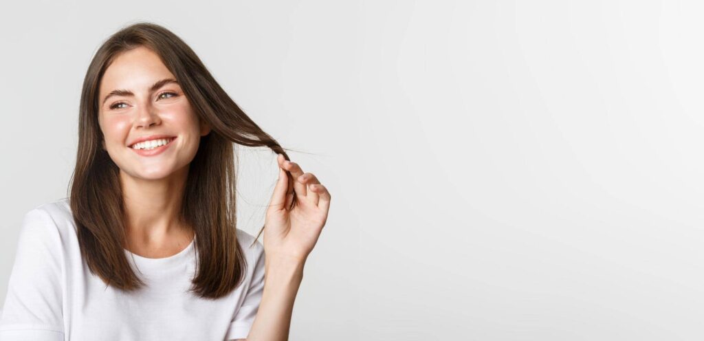 L'immagine raffigura una giovane donna con i capelli castani di media lunghezza, che sorride giocando con una ciocca dei suoi capelli fini. Indossa una maglietta bianca e sembra felice e rilassata, probabilmente soddisfatta dall’acquisto di uno shampoo per capelli fini, in posa contro uno sfondo chiaro e uniforme che mette in risalto il suo viso luminoso e l'espressione amichevole. Il suo sorriso genuino e il contatto visivo diretto con la camera creano un'atmosfera accogliente e positiva.