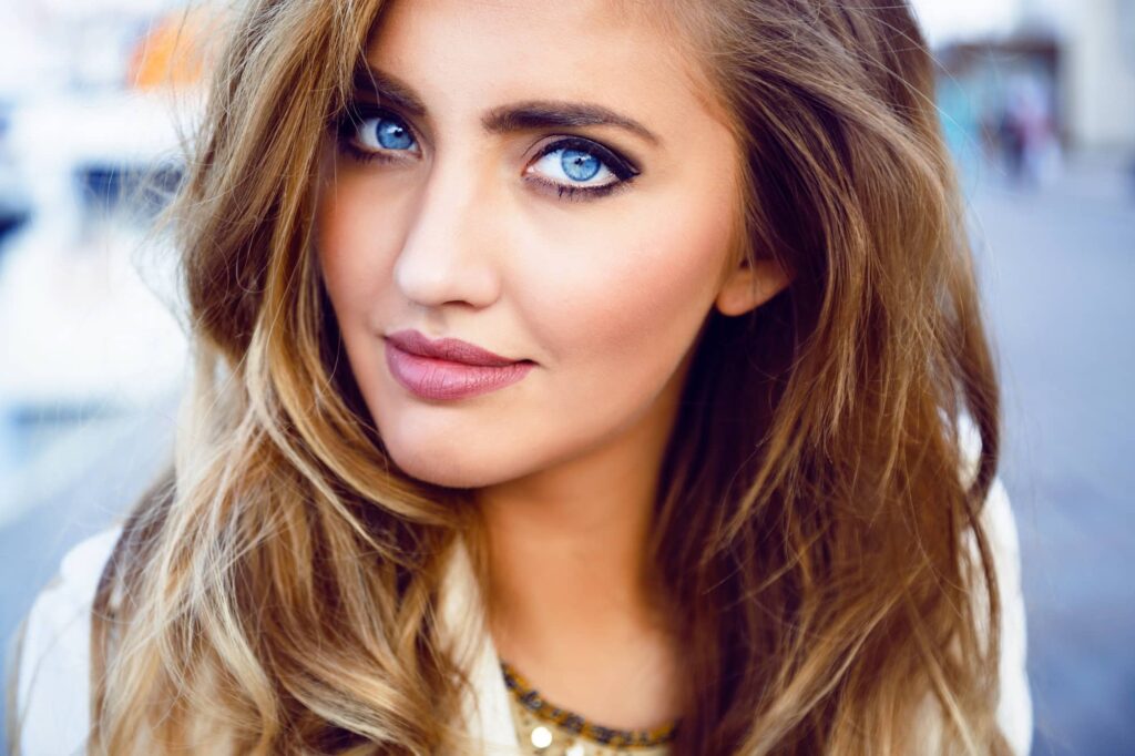 Primo piano di una giovane donna attraente con grandi occhi azzurri e labbra carnose. La sua pelle appare liscia e perfetta, e indossa un trucco naturale che enfatizza la sua bellezza. Ha i lunghi capelli acconciati in morbide onde, che aggiungono un tocco di eleganza al suo aspetto, mentre il castano chiaro è un perfetto colore di capelli per valorizzare gli occhi azzurri. Indossa anche una collana sottile, che completa il suo stile sofisticato.