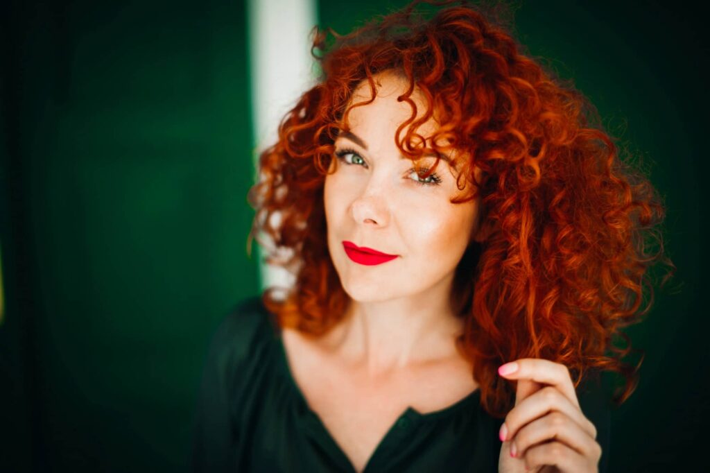 Donna dai capelli rossi ricci e vivaci e gli occhi verdi, che porta un rossetto rosso brillante e ha un'espressione pensierosa. Indossa un vestito scuro e sembra che stia toccando delicatamente una ciocca dei suoi capelli. Lo sfondo è di un verde uniforme che fa risaltare la tonalità dei suoi capelli e il trucco. Ha scelto un colore dei capelli per valorizzare gli occhi verdi.