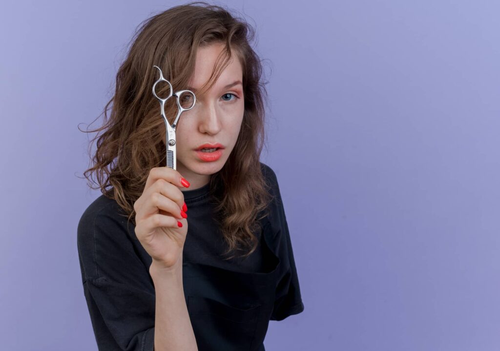 Ragazza con capelli castani ondulati di media lunghezza che tiene in mano delle forbici da parrucchiere e guarda attraverso uno degli anelli dell’impugnatura.
