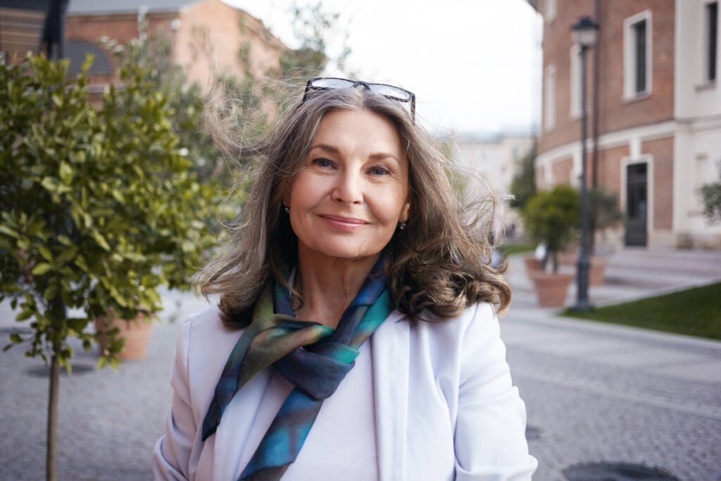 Sorridente donna matura con lunghi capelli grigi mossi dal vento e occhiali da vista in cima alla testa con sullo sfondo un paesaggio residenziale. Ha scelto un perfetto taglio di capelli per i 50 anni che la faccia apparire più giovane e fresca.