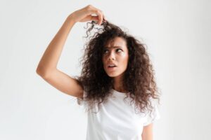 Ragazza con folti capelli lunghi e mossi tiene sollevata una ciocca con espressione frustrata, probabilmente chiedendosi quale potrebbe essere il miglior taglio per capelli folti che si adatti alla sua personalità e al suo stile di vita.