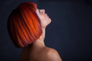 Una persona con una vivace colore di capelli che sfuma dal rosso all'arancione, sfoggia un taglio di capelli corto e moderno. La persona è di profilo e guarda verso l'alto su uno sfondo scuro.