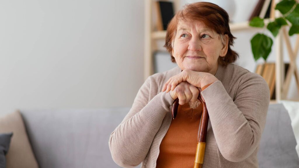 Donna anziana seduta con il mento e le mani appoggiate ad un bastone. Ha i capelli tinti di un colore scuro che potrebbe rientrare tra i colori di capelli che invecchiano.