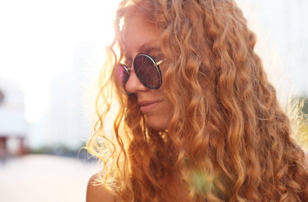Ragazza con lunghi capelli rossi ondulati e occhiali da sole che guarda di lato con la luce del sole alle spalle in uno spazio aperto. La ragazza è riuscita ad acconciare i capelli mossi in maniera impeccabile.