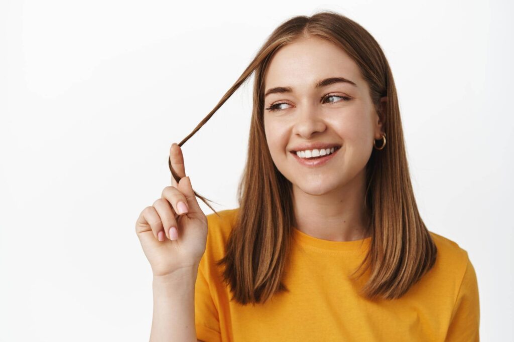 Ragazza sorridente che gioca con una ciocca dei suoi lunghi capelli fini. Potrebbe essere felice perché ha trovato il miglior assortimento di prodotti per capelli fini per dare volume alla sua chioma.