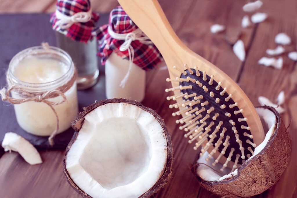 Immagine che mostra un insieme di prodotti per la cura dei capelli a base di cocco. In primo piano c'è una spazzola per capelli in legno con setole robuste su una mezza noce di cocco aperta. Accanto alla noce di cocco ci sono un barattolo di vetro con un balsamo per capelli fatto in casa a base di cocco e una bottiglia di vetro con quello che potrebbe essere olio di cocco, entrambi legati con un tessuto a quadri e spago per un aspetto rustico e naturale. Piccoli pezzi di cocco sono sparsi intorno, il tutto su un piano di legno che evoca un'atmosfera calda e organica. Questa composizione suggerisce un approccio naturale e nutriente alla cura dei capelli.