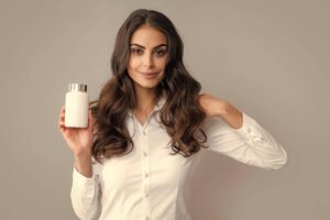 Un'immagine di una donna con capelli lunghi e ondulati che indossa una camicia bianca e tiene in mano una bottiglia di balsamo per capelli. Il suo sguardo diretto e un lieve sorriso possono indicare fiducia nel prodotto che sta presentando. Il contesto neutro enfatizza sia la modella che la bottiglia, suggerendo un focus sulla cura e sulla salute dei capelli.