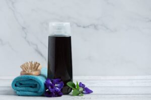 Un flacone di shampoo scuro con dispenser bianco è posizionato su un tavolo di legno bianco accanto a un asciugamano turchese arrotolato e una spazzola per capelli in legno con setole naturali. Fiori di colore viola intenso con foglie verdi aggiungono un tocco di colore e suggeriscono ingredienti botanici naturali, collegandosi all'importanza di scegliere shampoo con ingredienti di qualità. L'immagine comunica un senso di purezza e cura naturale per i capelli. Scopri lo shampoo per capelli ideale per te.