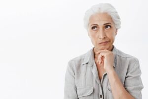 Una donna anziana con capelli bianchi in una camicia grigia guarda pensierosamente verso l'alto a sinistra, con la mano sotto il mento come se fosse immersa in profonde riflessioni, probabilmente cercando il miglior colore per coprire i capelli bianchi.