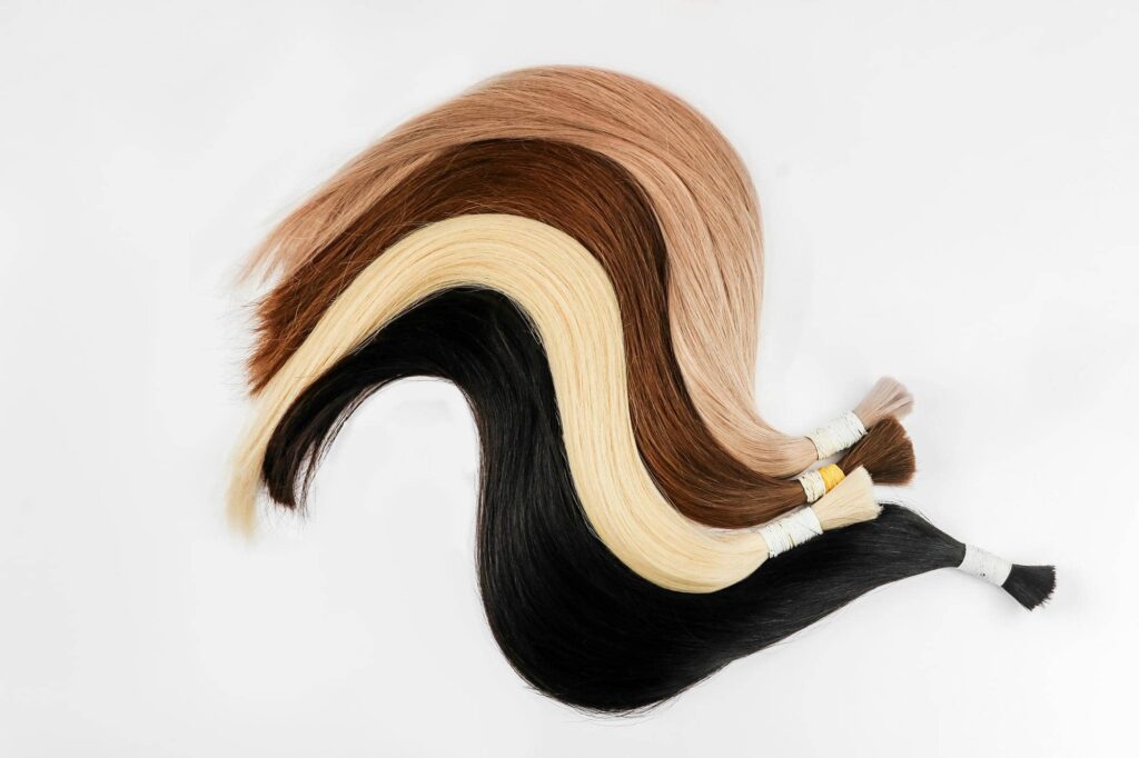 Una serie di ciocche di capelli di varie tonalità, che vanno dal nero, al biondo platino, al castano chiaro e scuro, disposte in modo ondulato su uno sfondo bianco. Ogni ciocca è legata con un nastro o elastico, suggerendo che si tratti di campioni di capelli per estensioni. Il contrasto tra i diversi colori di capelli e lo sfondo bianco risalta notevolmente, facendo emergere la varietà di tonalità disponibili. La disposizione dei campioni crea un effetto visivo armonioso e piacevole. Una volta applicate ai capelli, sarà importante evitare gli errori comuni con le extension per capelli.