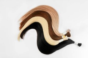 Una serie di ciocche di capelli di varie tonalità, che vanno dal nero, al biondo platino, al castano chiaro e scuro, disposte in modo ondulato su uno sfondo bianco. Ogni ciocca è legata con un nastro o elastico, suggerendo che si tratti di campioni di capelli per estensioni. Il contrasto tra i diversi colori di capelli e lo sfondo bianco risalta notevolmente, facendo emergere la varietà di tonalità disponibili. La disposizione dei campioni crea un effetto visivo armonioso e piacevole. Una volta applicate ai capelli, sarà importante evitare gli errori comuni con le extension per capelli.