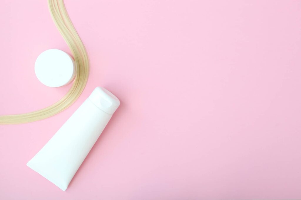 Una composizione minimalista con una ciocca di capelli biondi disposta in modo da creare una curva morbida su uno sfondo rosa pastello. Inoltre, c'è una bottiglia bianca, probabilmente di balsamo, e un barattolo rotondo con coperchio, che potrebbe contenere un prodotto per la cura dei capelli come una maschera. L'intera scena ha un aspetto pulito e ordinato, con balsamo e maschera per capelli che, uniti alla ciocca di capelli, comunicano un messaggio di bellezza e cura personale.