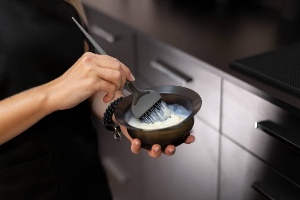 Una persona mescola la tinta per capelli in una ciotola con un pennello applicatore, preparandosi per un trattamento di colorazione. Scopri le differenze tra shampoo colorato e tinta per capelli.