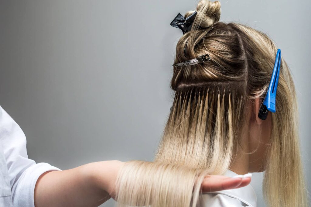 Immagine che mostra il processo di applicazione delle extension per capelli fini in un salone di bellezza. È visibile il profilo di una cliente con i capelli biondi, parzialmente raccolti e fermati con clip di plastica nere e blu. Una sezione di capelli è stata separata per mostrare le estensioni già applicate alla radice, che si distinguono per il loro colore più scuro rispetto al resto dei capelli. Le estensioni sono attaccate vicino al cuoio capelluto e cadono liberamente con i capelli naturali della cliente. La mano di un parrucchiere in una camicia bianca solleva delicatamente la sezione di capelli con le estensioni, indicando un lavoro di precisione.