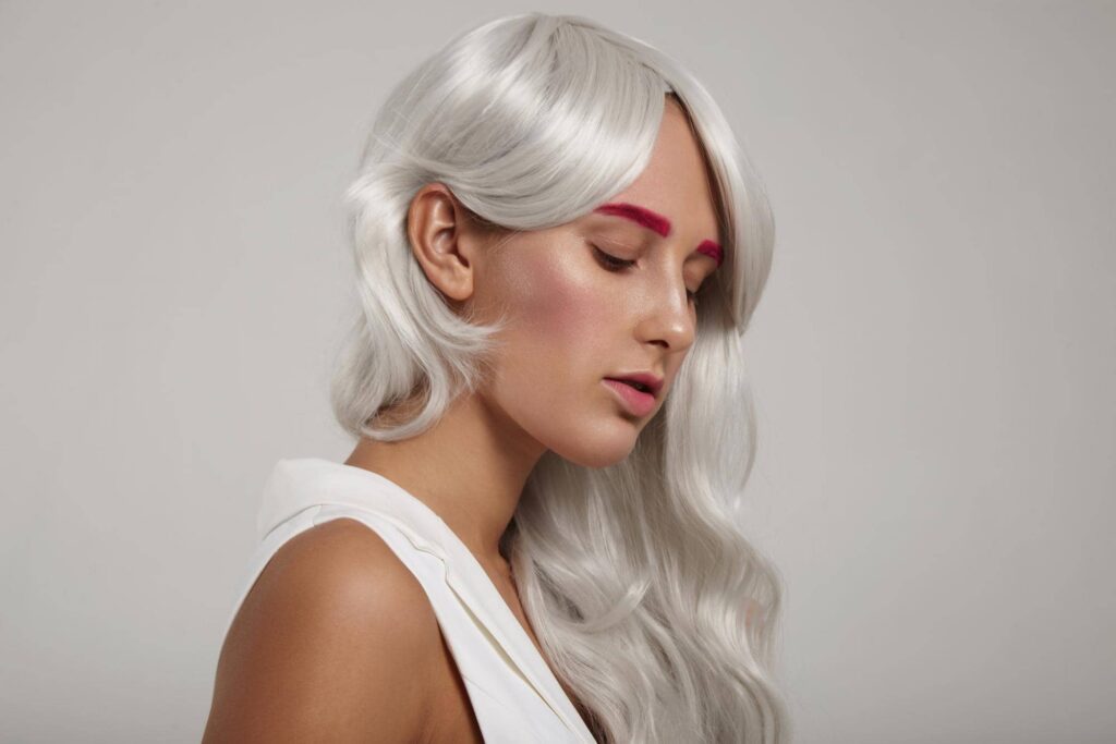 Ritratto di una donna che è riuscita ad ottenere i capelli grigi lussuosi e ben curati, acconciati con morbide onde. Sfoggia sopracciglia audaci colorate di rosa che contrastano con la tonalità platino dei capelli, evidenziando un look moderno ed elegante che rispecchia le ultime tendenze.
