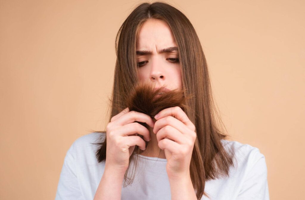Una donna con espressione preoccupata che esamina attentamente le punte dei suoi capelli. I capelli sembrano danneggiati o spezzati, il che potrebbe essere motivo della sua preoccupazione. Indossa una semplice maglietta bianca e si trova su uno sfondo beige neutro, il che fa risaltare maggiormente il suo volto e i capelli. La scena cattura un momento di cura personale con un focus sui problemi legati alla salute dei capelli, risolvibili con una maschera per capelli alla cheratina.