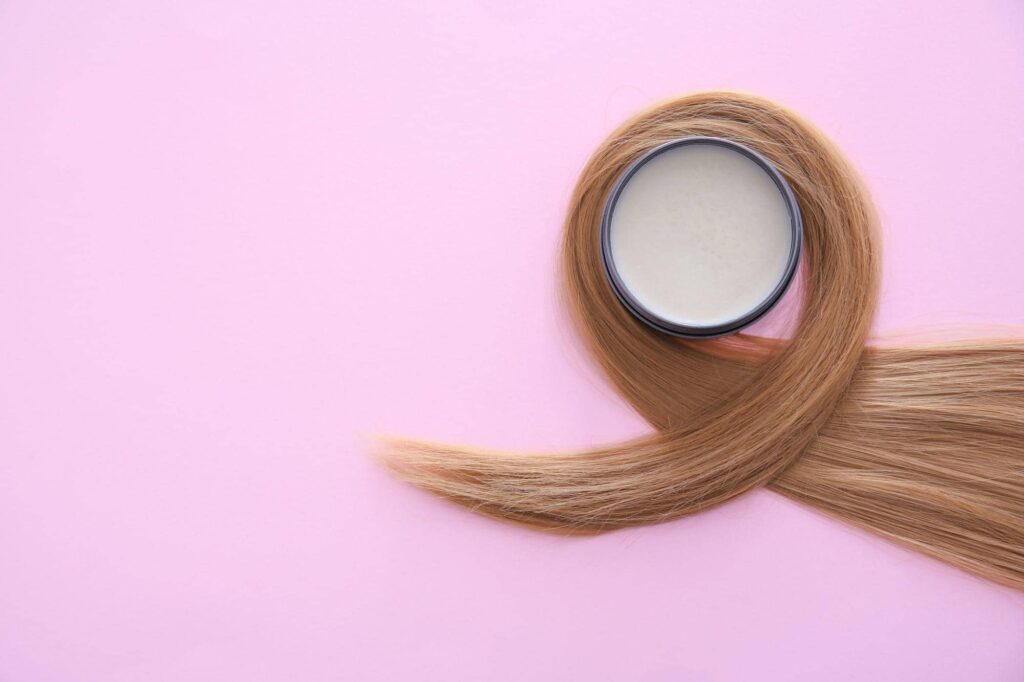 Un vasetto di maschera ristrutturante per capelli è avvolto da una ciocca di capelli biondi su uno sfondo rosa chiaro. Questa immagine rappresenta il concetto di cura e styling dei capelli, evidenziando l'importanza della cura e della ristrutturazione per ottenere una chioma sana e ben curata.