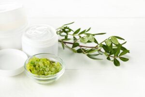 Una disposizione di prodotti per la cura di capelli e del corpo su un tavolo con sfondo bianco. In primo piano, c'è una ciotola di vetro trasparente contenente cristalli di sale verde, presumibilmente sale da bagno o esfoliante. Dietro la ciotola di sale, si trovano due barattoli di maschere per capelli naturali, uno aperto con il contenuto visibile e l’altro chiuso. Sul lato destro dell'immagine c'è un ramo di una pianta verde con foglie piccole, che aggiunge un tocco naturale e fresco alla composizione. Questi elementi insieme suggeriscono un'atmosfera di spa o relax domestico, enfatizzando la naturalezza e la cura personale.
