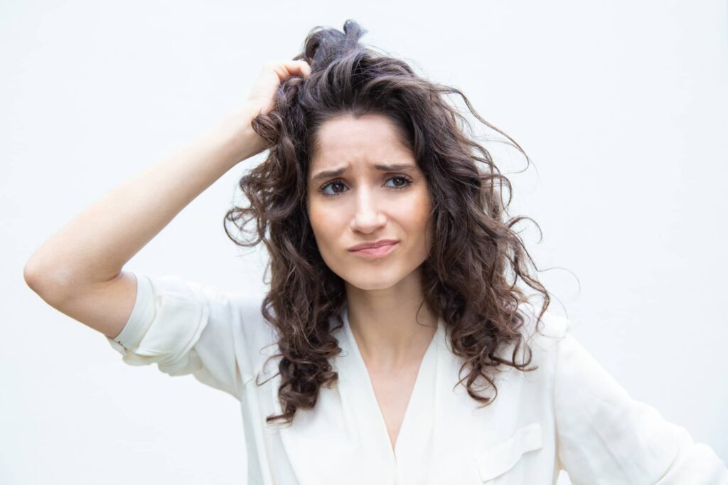 Un'immagine di una donna con espressione preoccupata e confusa, grattandosi la testa mentre guarda verso l'obiettivo. I suoi capelli ricci sembrano disordinati e unti, il che potrebbe suggerire che lei sta considerando i migliori shampoo per capelli grassi a sua disposizione.