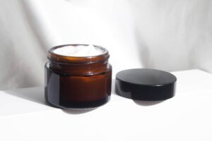 Immagine che mostra un barattolo di vetro marrone traslucido con una crema bianca all'interno, probabilmente una delle migliori maschere per capelli, e il suo coperchio nero posto accanto. Il contenitore è posizionato su una superficie bianca con uno sfondo che sembra essere un tessuto leggero e delicato. L'illuminazione è morbida e diffusa, suggerendo che la foto potrebbe essere stata scattata alla luce naturale.