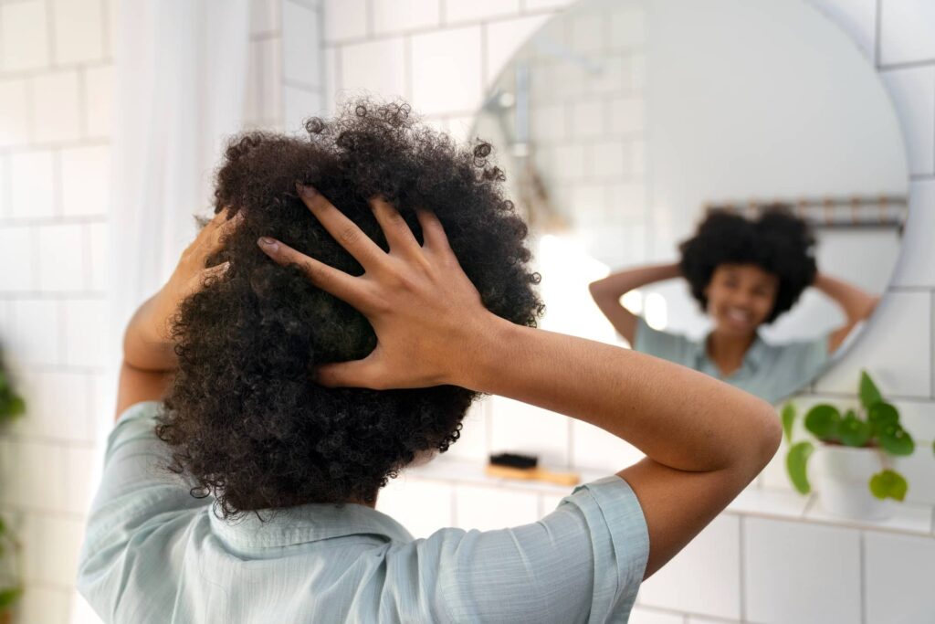 Ragazza di colore che si guarda allo specchio in bagno mentre si passa le mani tra i ricci e crespi capelli scuri, forse pensando a quali prodotti per capelli crespi potrebbe utilizzare per rendere la sua chioma più gestibile.