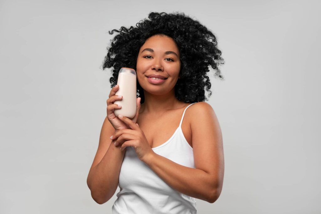 Immagine che mostra una giovane donna sorridente che tiene una bottiglia di balsamo per capelli vicino al suo viso, probabilmente uno dei migliori balsami per capelli ricci. La donna ha capelli ricci e voluminosi e indossa una canotta bianca. Lo sfondo è di colore chiaro e uniforme, il che mette in evidenza il soggetto e il prodotto che sta presentando. Il suo sorriso accogliente e l'atteggiamento rilassato trasmettono un'immagine positiva in un contesto di promozione della salute dei capelli.