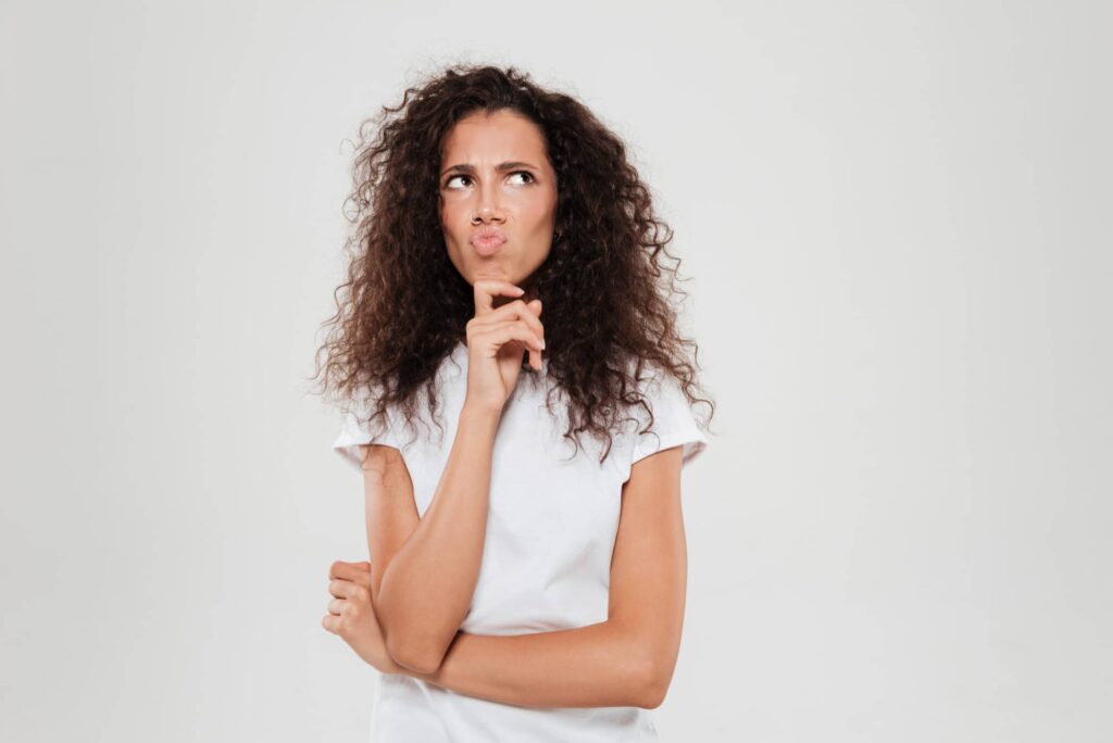 Una donna con capelli ricci e crespi sta pensando intensamente con una mano sul mento e lo sguardo distratto verso l'alto a sinistra, mostrando un'espressione interrogativa, probabilmente mentre si chiede quali siano le migliori maschere per capelli crespi. Questa immagine potrebbe rappresentare la comune preoccupazione riguardo alla gestione dei capelli crespi.