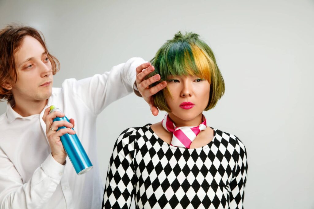 Parrucchiere esperto che utilizza uno spray per capelli sulla chioma dai colori verde-gialli di una ragazza asiatica.