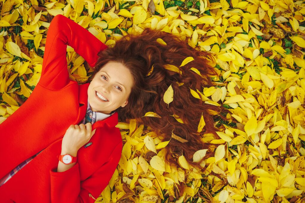 Ragazza con lunghi capelli ondulati castano-rossastri e un cappotto rosso distesa su un letto di foglie gialle in un ambiente autunnale. Questa stagione non è il momento ideale per tagliare i capelli.