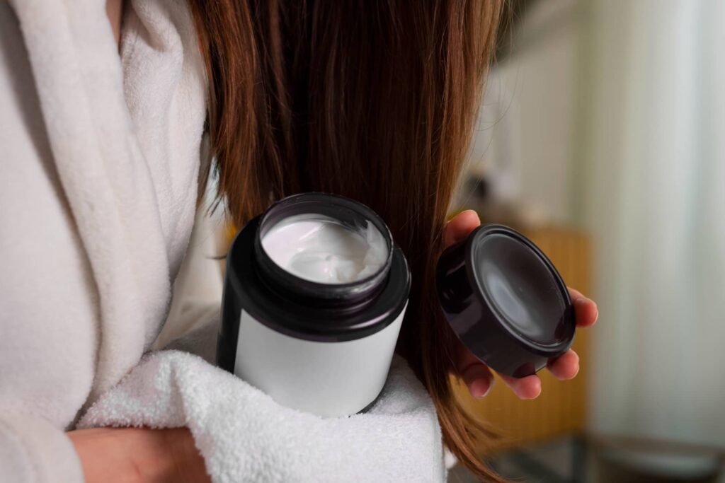 Una donna con un accappatoio bianco tiene aperto un barattolo di balsamo per capelli, pronto per essere utilizzato nella sua routine di cura dei capelli. I lunghi e lisci capelli castani della donna pendono liberamente in attesa del trattamento idratante. L’immagine è la rappresentazione ideale del quando e come usare il balsamo per capelli.
