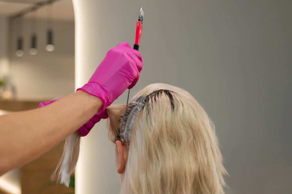 Una parrucchiera professionista che indossa guanti rosa e applica un decolorante alla ricrescita di capelli tinti di un'altra persona. La persona i cui capelli vengono trattati è vista da dietro e non è possibile vedere il suo viso. L'attenzione è focalizzata sull'applicazione del prodotto sui capelli, utile per prolungare la durata di una tinta per capelli.