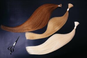 Tre diverse tonalità di extension per capelli, variando dal castano al biondo chiarissimo, sono disposte elegantemente su uno sfondo scuro. Le ciocche di capelli veri mostrano una transizione di colore liscia e naturale, ideali per esaltare qualsiasi acconciatura. Accanto alle extension, un paio di forbici professionali per parrucchiere giace sullo sfondo. L'immagine enfatizza la bellezza e la varietà delle opzioni disponibili per le extension, ognuna delle quali presenta una diversa durata delle extension ai capelli.