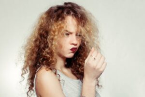 Foto di una giovane donna con lunghi capelli ricci, che guarda verso con un'espressione un po' frustrata una ciocca di capelli crespi che tiene tra le dita, probabilmente pensando alla sua necessità di uno shampoo anticrespo. I suoi capelli hanno diverse sfumature di biondo e castano, che enfatizzano la texture riccia. Lei indossa un top senza maniche grigio chiaro, il trucco è naturale con l'eccezione di un rossetto scuro che dà un contrasto forte con il resto dei toni neutri dell'immagine. Lo sfondo è di colore grigio.