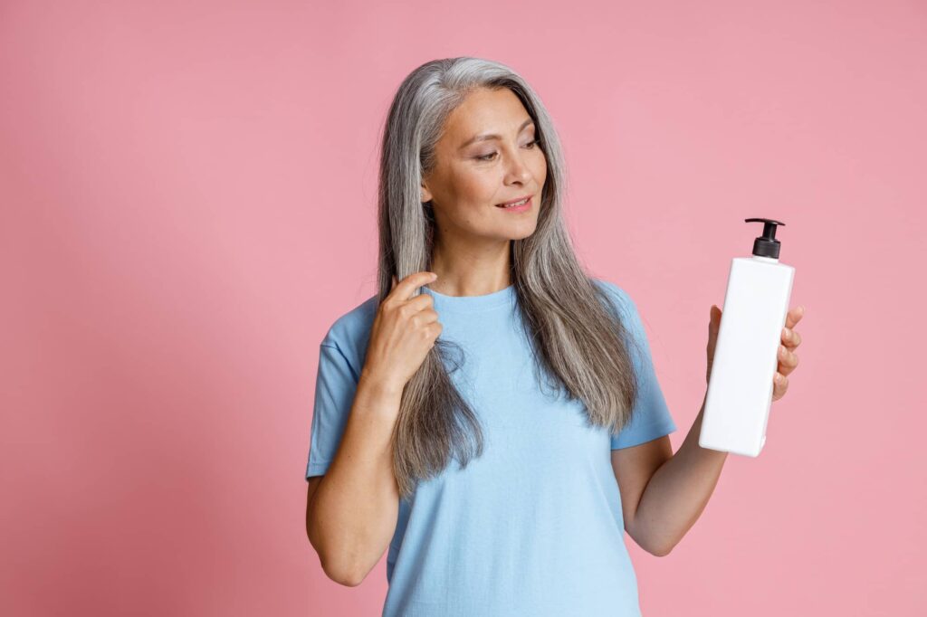 Immagine che mostra una donna matura con i capelli grigi lunghi e lisci che guarda dolcemente verso il lato mentre tiene una bottiglia di shampoo antigiallo con erogatore nero. Indossa una maglietta azzurra ed è posizionata contro uno sfondo rosa che crea un piacevole contrasto con il suo abbigliamento e i suoi capelli. La donna contempla il prodotto nella sua mano, forse pensando alle sue proprietà o come applicarlo, suggerendo un focus sulla cura personale.