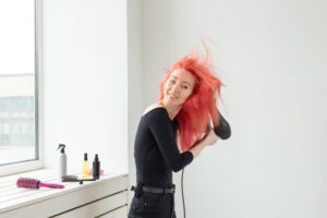 Immagine che mostra una donna con vivaci capelli rossi mentre si asciuga i capelli con un phon. Ha un'espressione divertita e rilassata sul viso e indossa una maglietta nera a maniche lunghe e pantaloni neri. Sul davanzale della finestra accanto a lei si trovano alcuni prodotti per la cura dei capelli, inclusi flaconi spray, oli, spazzole e shampoo per capelli colorati. La stanza è luminosa, con luce naturale che entra dalla finestra, e lo sfondo è una parete bianca che contrasta con il colore brillante dei suoi capelli.