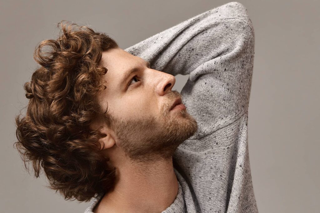 Vista laterale di un ragazzo con lunghi capelli ricci castano chiaro che guarda verso l’alto con un braccio dietro la testa. Il suo taglio di capelli da uomo riflette uno stile sbarazzino.