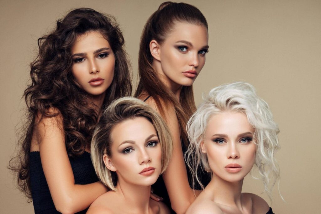 Quattro ragazze con diverse capigliature che guardano in camera, disposte in due file ad altezze diverse. Le capigliature sono: capelli lunghi, scuri e ondulati; capelli lunghi, castani e lisci; capelli medio-corti, scuri alla radice e poi biondo cenere, lisci; capelli medio-lunghi, bianchi e ondulati.