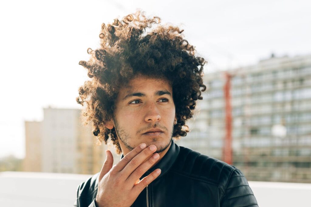 Ragazzo pensieroso con voluminosi capelli ricci castani mossi dal vento in un contesto urbano. Il suo taglio di capelli da uomo valorizza al meglio la texture naturale della sua chioma.