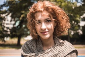 Ragazza sorridente con capelli mossi di colore rossastro e lunghezza media mossi dal vento con un parco alberato sullo sfondo. Porta un taglio per capelli mossi che valorizza al meglio la sua figura.