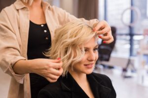 Foto di una parrucchiera che con mani esperte acconcia i capelli mossi di media lunghezza di una donna bionda.
