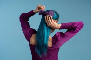 Donna che dà le spalle alla telecamera mentre si sistema un fiocco viola tra i lunghi e lisci capelli color turchese.