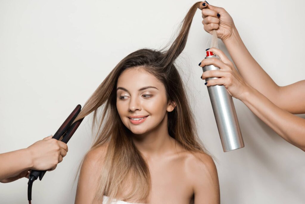 Ragazza sorridente che lascia che due parrucchiere si prendano cura della sua chioma con una piastra per capelli e uno spray lisciante, uno dei più comuni metodi per lisciare i capelli.